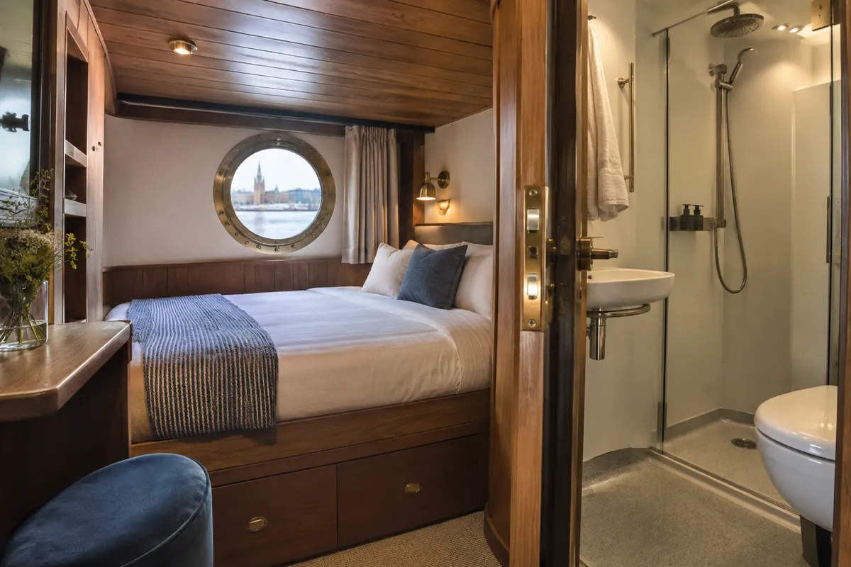 Compact ship cabin with a low ceiling, porthole window, double bed, and en-suite bathroom